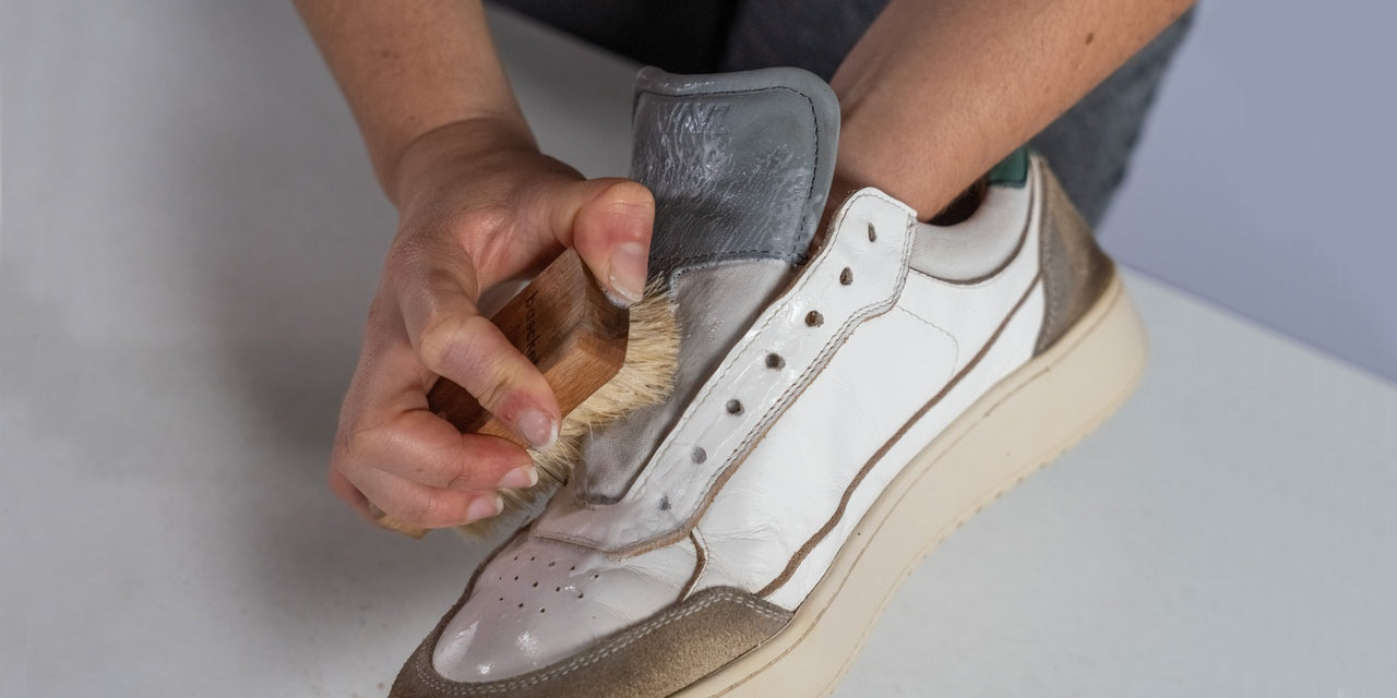 the tongue of a pair of the hedonist white grey green sneakers being cleaned with a brush