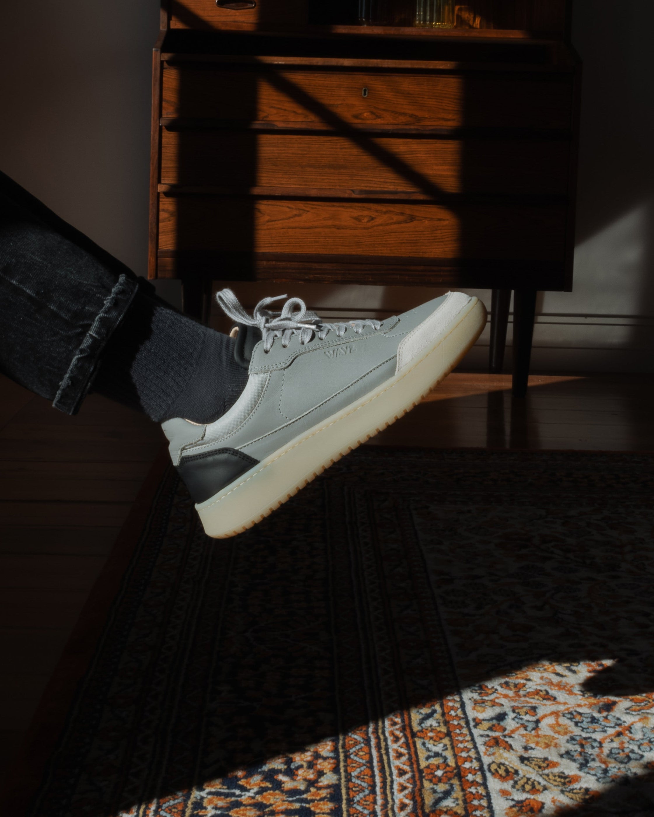 Person wearing gray Hedonist sneakers with gum soles on a dark wooden floor.