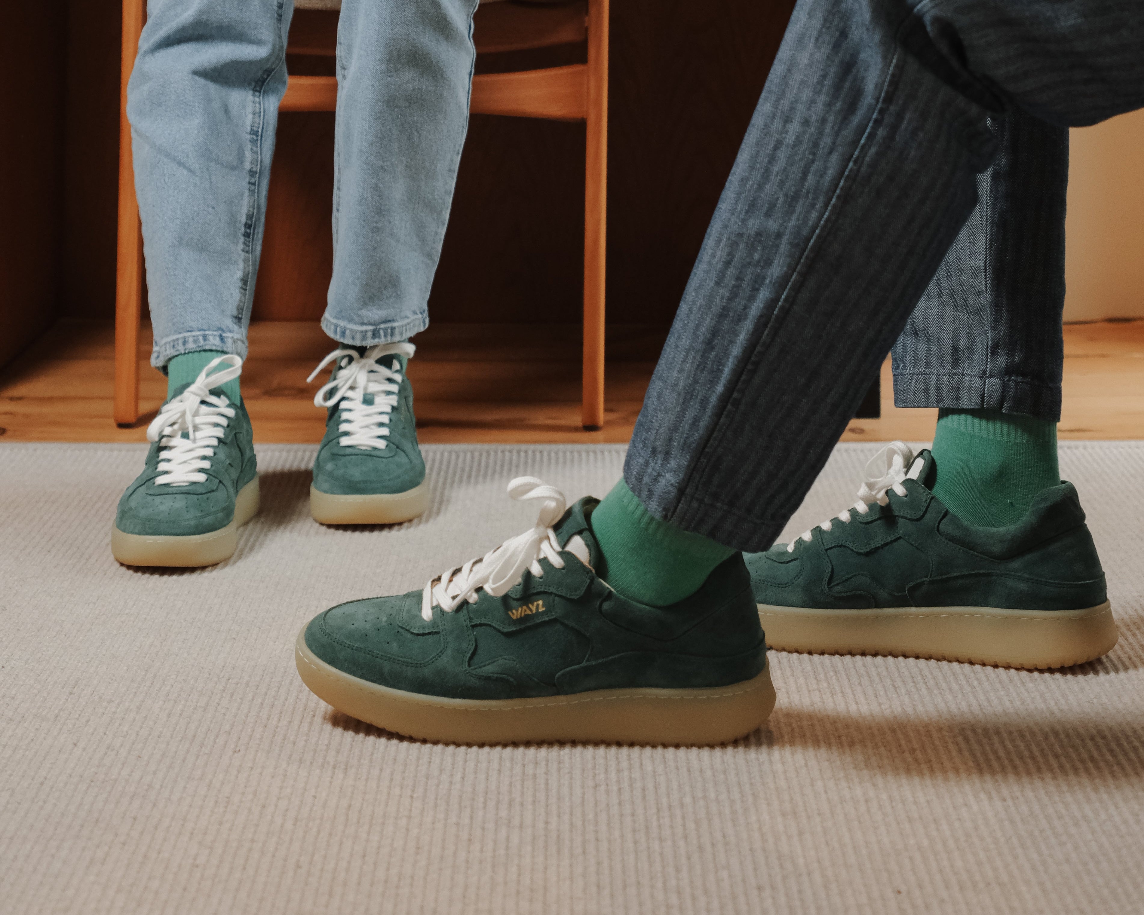 Two people wearing pine green suede sneakers in a room with wooden furniture.
